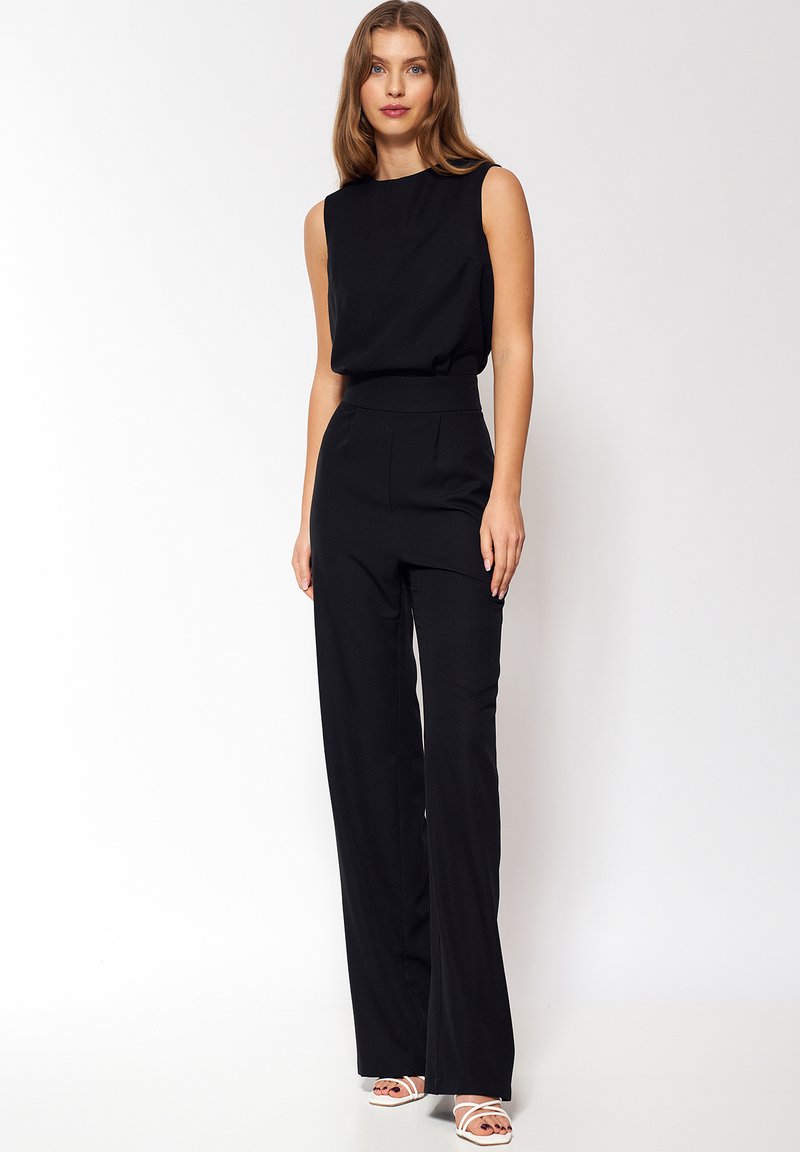 Czarny jednoczęściowy strój bez rękawów z dopasowanym pasem i szerokimi nogawkami. Gładki materiał bez wzorów. Białe sandały na paski.