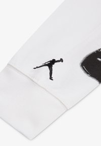 Biała rękaw od bluzy z kapturem z czarnym haftowanym logo Jumpman blisko ściągacza, o gładkiej fakturze i widocznych detalach szwów.