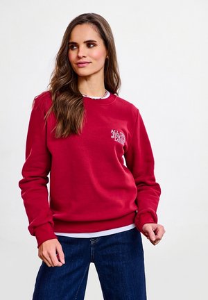Czerwona bluza z miękkiego materiału z prążkowanymi mankietami i dołem. Na piersi białe napisy, warstwowo noszona na białej koszuli, zestawiona z ciemnymi dżinsami.
