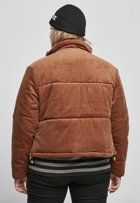 Bruine corduroy pufferjack met horizontale stiksels, hoge kraag en gouden hardware accenten. Draag het over een gestreept topje met zwarte broek.