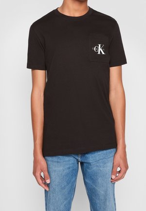 Mand iført en sort t-shirt med korte ærmer og en Calvin Klein-logo på brystlommen samt lyseblå jeans.