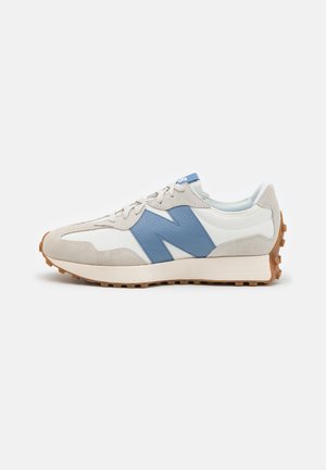 Zapatilla blanca y beige con un gran logo azul "N", detalles en ante beige, cordones blancos y suela de goma marrón texturizada.