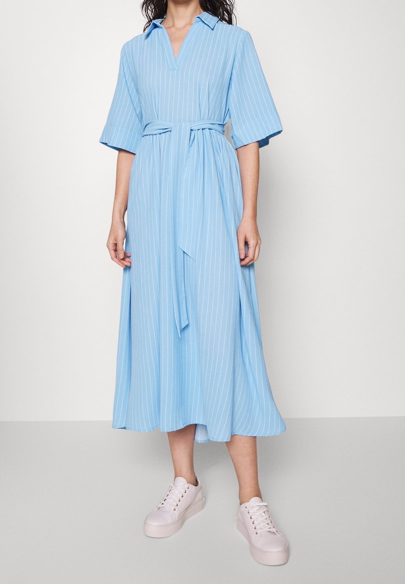 Ljusblå randig klänning med korta ärmar, V-hals och knytmidja. Tillverkad av lättviktsmaterial, når till kycklingbenet, bärs med vita sneakers.