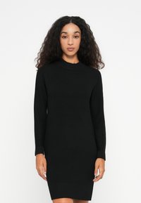 Una donna sta in piedi di fronte, indossando un vestito nero a maglia con maniche lunghe e lunghezza al ginocchio, su uno sfondo bianco semplice.