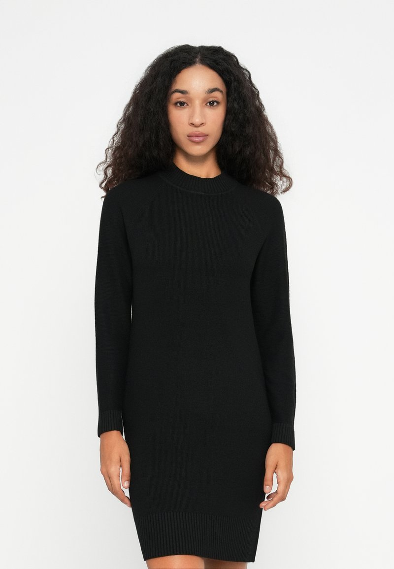 Una donna sta in piedi di fronte, indossando un vestito nero a maglia con maniche lunghe e lunghezza al ginocchio, su uno sfondo bianco semplice.