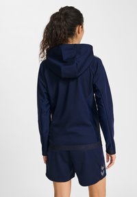 Marinblå atletisk hoodie med huva, långa ärmar och strukturerat tyg; har reflekterande detaljer och en logotyp vid midjan.