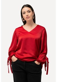 Blusa in satin rosso con scollo a V, maniche a palloncino e dettagli legati ai polsini. Abbinata a pantaloni neri, che mostrano una texture liscia.