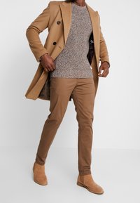 Cappotto in misto lana color tan con bottoni doppi, maglione marrone testurizzato, pantaloni beige e stivali alla caviglia in suede marrone.