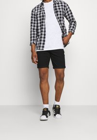 Camisa de cuadros en blanco y negro sobre una camiseta blanca, pantalones cortos negros enrollados y zapatillas negras con detalles blancos, de pie sobre un fondo liso.
