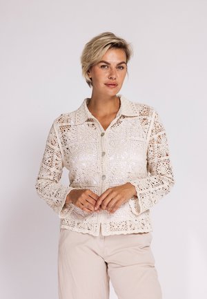 Woman with short blonde hair wearing a beige crochet button-up blouse and light beige pants, standing against a plain white background.