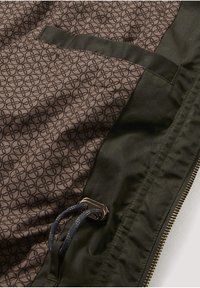 Dunkelgrünes Jackenfutter mit gemustertem braunem Stoff, einer Innentasche, einem Kordelzug mit Metallstopp und einem schwarzen Reißverschluss entlang des Rands.