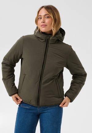 Femme portant une doudoune à capuche vert olive et un jean bleu, debout les mains touchant les côtés de la veste, fond neutre.