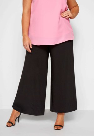 Schwarze weit geschnittene Hosen mit glatter Textur, kombiniert mit einem hellrosa ärmellosen Oberteil. Das Outfit wird mit schwarzen, geschnürten High Heels abgerundet.