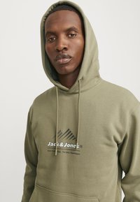 Olive green hoodie with a drawstring, featuring a logo and mountain graphic. Made of soft fabric with a standard fit and kangaroo pocket.