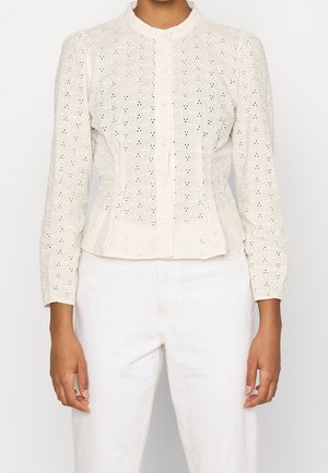 Blouse blanche en œillet avec manches longues, col rond et fermeture à boutons sur le devant. Le tissu présente des motifs découpés complexes et une coupe ajustée.