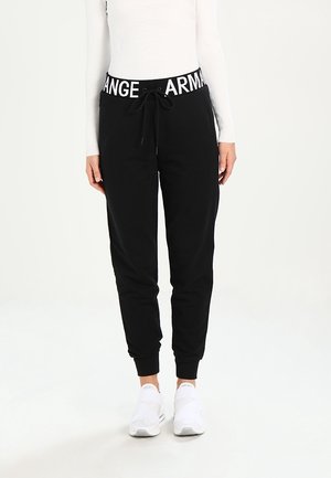 Personne portant un pantalon de jogging noir avec une ceinture blanche siglée Armani Exchange, un haut blanc à manches longues et des baskets blanches, debout sur un fond blanc.