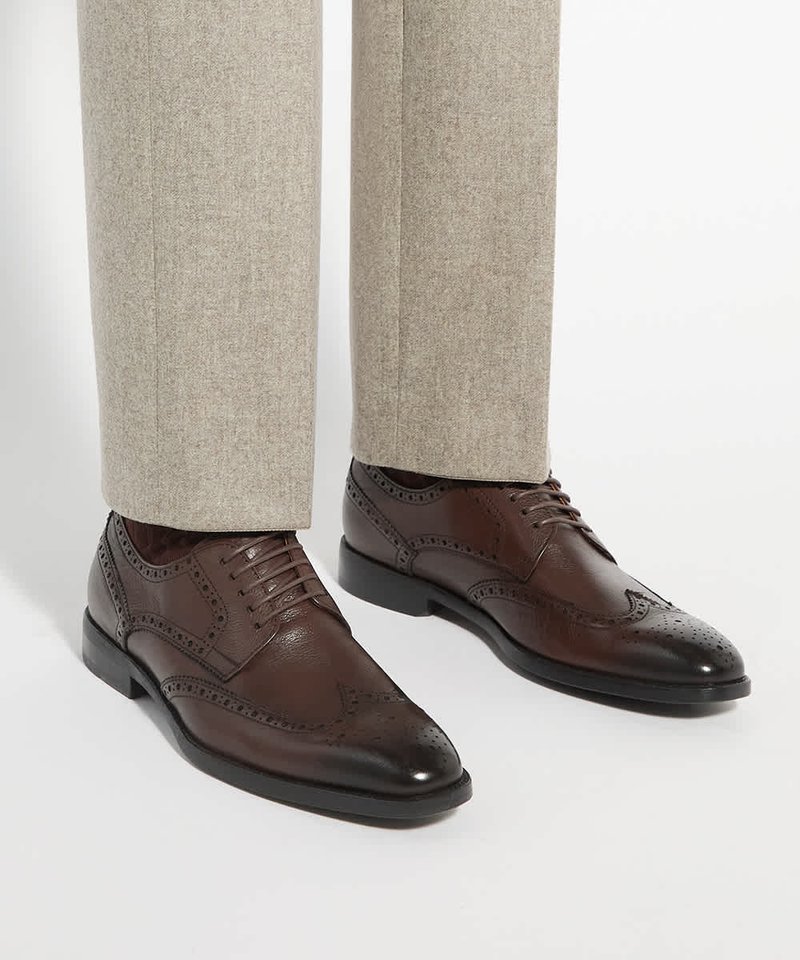 Chaussures brogue en cuir marron portées avec un pantalon habillé beige clair sur un fond blanc uni.