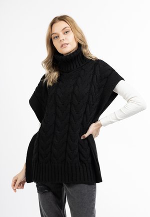 Schwarzer Kabelstrick-Poncho mit hohem Kragen, versehen mit einem gerippten Saum und seitlichen Schlitzen, getragen über einem langärmeligen weißen Hemd und zerfetzten grauen Jeans.