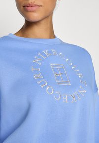 Lyseblå hettegenser laget av mykt stoff med en sirkulær, brodert Nike Court-logo i kremfarge. Løs passform med rund hals.