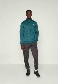 Sarkans zip-up jaka ar melnu akcentiem un logo, saskaņots ar pelēkiem biksēm. Modelis valkā melnus sporta apavus, stāvot uz neitrāla fona.