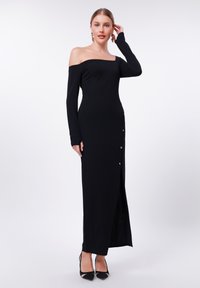 Svart off-the-shoulder kjole med lange ermer, pyntet med en frontsplitt og gullknapper på den ene siden. Glatt, elastisk stoff.