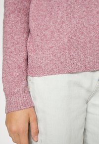 Różowy, teksturowany sweter z dzianiny z prążkowanymi mankietami, noszony na jasnych dżinsach. Zbliżenie pokazujące szczegóły rękawa i dłoni.