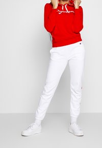 Czerwony hoodie z białymi sznurkami i logo, w połączeniu z białymi dresami z mankietami na kostkach i akcentami logo, noszony z białymi trampkami.
