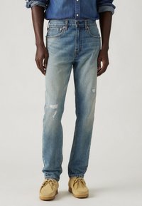 Blugi din denim albastru deschis cu un croi drept, având detalii distresate și un design clasic cu cinci buzunare, asortați cu pantofi mocasini bej.