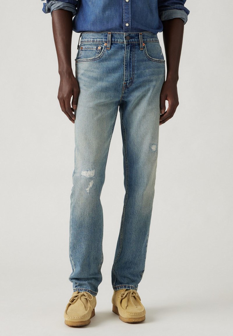 Blugi din denim albastru deschis cu un croi drept, având detalii distresate și un design clasic cu cinci buzunare, asortați cu pantofi mocasini bej.