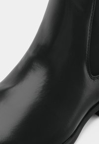 Nahaufnahme eines glänzenden schwarzen Leder-Chelsea-Boots, der die glatte Textur und das elastische Seitenpanel auf einem weißen Hintergrund zeigt.