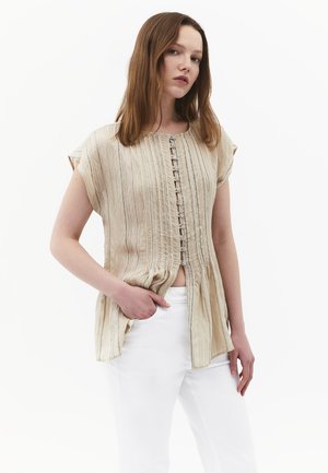 Vrouw met steil bruin haar die een beige korte mouw met knopen en een witte broek draagt, poseert met één hand in haar zak.
