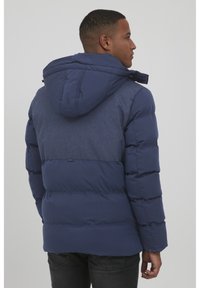 Navy puffer jacket with a hood, featuring a textured upper panel, quilted body, and side pockets. Subtle logo detail below the back yoke.