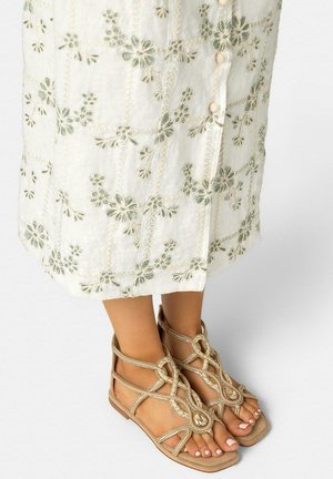 Pies con sandalias planas beige de tiras con detalles trenzados, junto a una falda blanca con bordado floral verde y botones decorativos.