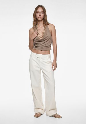 Femme portant un haut taupe sans manches drapé, des colliers en or superposés, un pantalon blanc à jambes larges, et des sandales beige avec des ornements.
