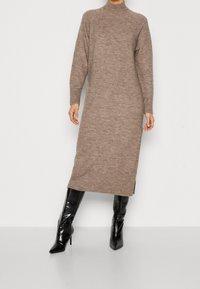 Robe en maille col roulé dans une teinte marron douce avec des poignets côtelés et des fentes latérales, associée à des bottes noires brillantes montant jusqu'aux genoux.