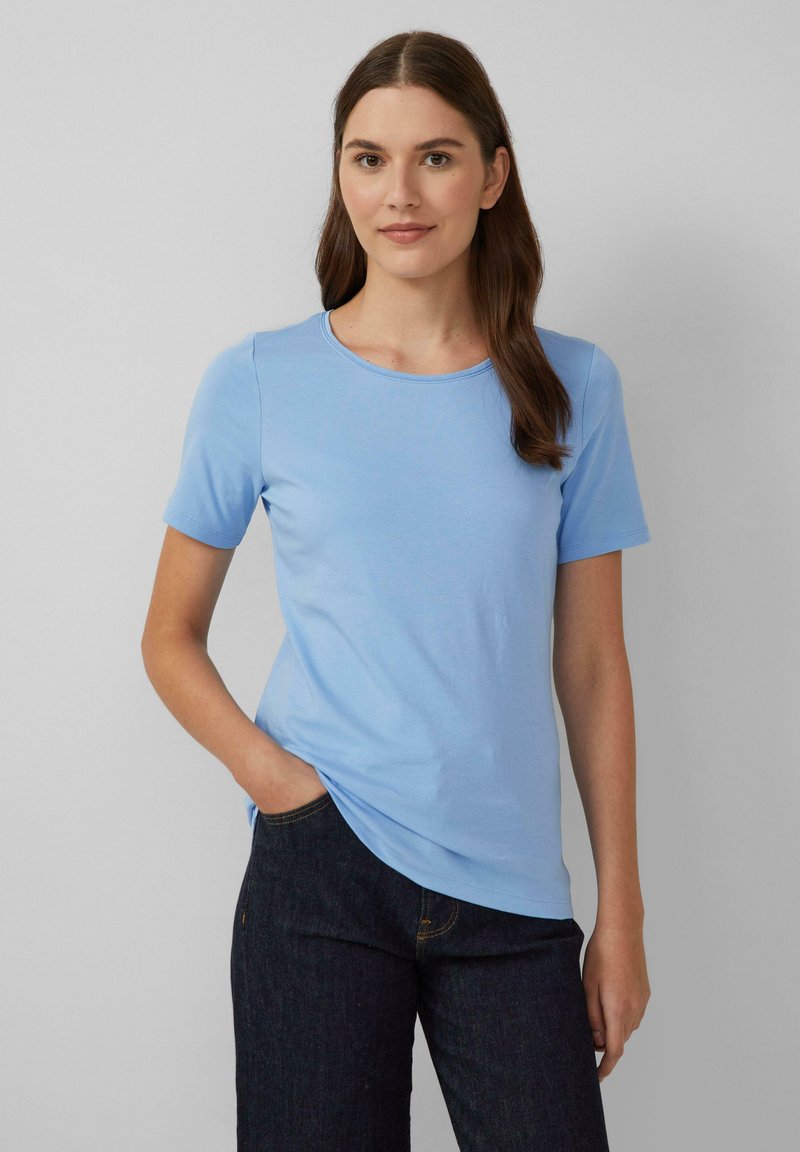 Vrouw met lang bruin haar in een lichtblauwe T-shirt met korte mouwen en donkere spijkerbroek, staand met één hand in haar zak tegen een grijze achtergrond.
