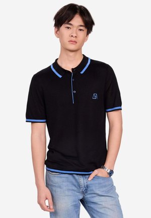 Junger Mann trägt ein schwarzes Poloshirt mit blauem Besatz und hellblauen Jeans, steht mit einer Hand in der Tasche vor weißem Hintergrund.