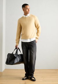 Beige Strickpullover, darunter ein weißes Hemd mit Kragen, dunkle Jeans, schwarze Ledertasche mit abgerundeten Kanten und doppelten Griffen, flache Schuhe mit Akzenten.