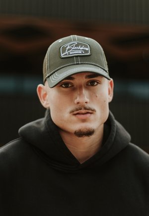 Green baseball cap with a white embroidered logo, worn with a black hooded sweatshirt. Background is blurred with dark tones.