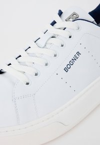 Fehér bőr sneaker, sötétkék részletekkel, hat fűzőlyukkal, lapos fehér fűzőkkel, domború márkanevével és perforációkkal az oldalon.