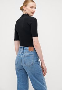 Μαύρο κοντομάνικο κορμάκι συνδυασμένο με ψηλόμεσα ανοιχτό μπλε τζιν, που διαθέτει δερμάτινη ετικέτα μάρκας και δύο πίσω τσέπες.