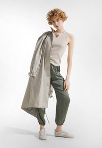 Mujer con cabello rizado que lleva una blusa sin mangas beige y un abrigo ligero tipo gabardina colgado sobre un hombro, pantalones jogger verde olivo y zapatillas deportivas beige.