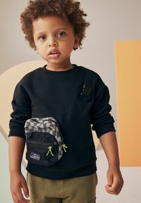 Svart sweatshirt med långa ärmar, kombinerad med olivgröna byxor. Liten, svart och grå rutig axelväska med neongröna detaljer.