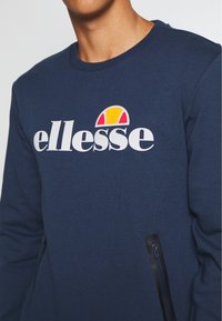 Sweat-shirt bleu marine avec un col rond et un logo "ellesse" blanc. Dispose de poches latérales zippées pour plus de fonctionnalité.