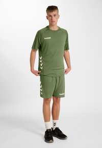 Groen sportoutfit bestaande uit een shirt met korte mouwen en een korte broek, met witte zijkanten en branding, gemaakt van gladde stof.
