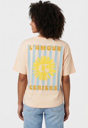 Persona con capelli ricci che indossa una maglietta beige con strisce blu, una grafica di un sole giallo e il testo "L'AMOUR CERISES" sul retro.