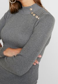 Harmaa ribbineulottu yläosa, jossa korkea kaulus, pitkät hihat ja koristeelliset kullanväriset napit olkapäällä, esitellen pehmeää materiaalia.