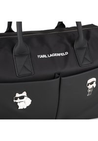 Geantă tote neagră cu logo-ul Karl Lagerfeld, având două ace de email decorative: o pisică și o figură, ambele purtând ochelari de soare. Textură netedă.