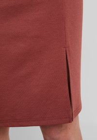 Jupe crayon couleur rouille avec une texture lisse, dotée d'une fente latérale et de coutures nettes le long de l'ourlet et de la couture.