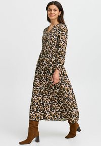 Långärmad maxiklänning med ett flerfärgat leopardmönster på en ljus bakgrund, med en rynkad midja och V-ringning. Bärs med knähöga bruna stövlar.